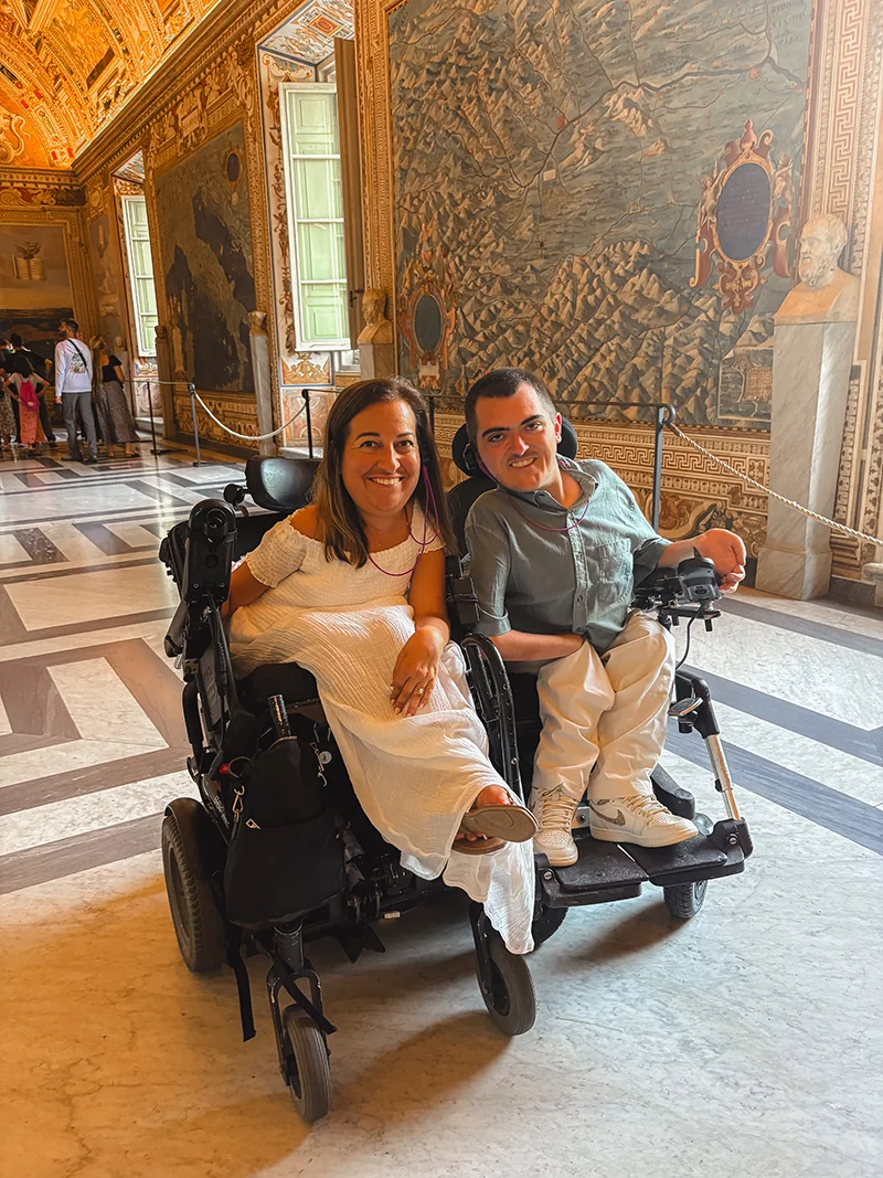 A woman in a wheelchair wearing a white dress with a man in a wheelchair who are smiling in a hallway