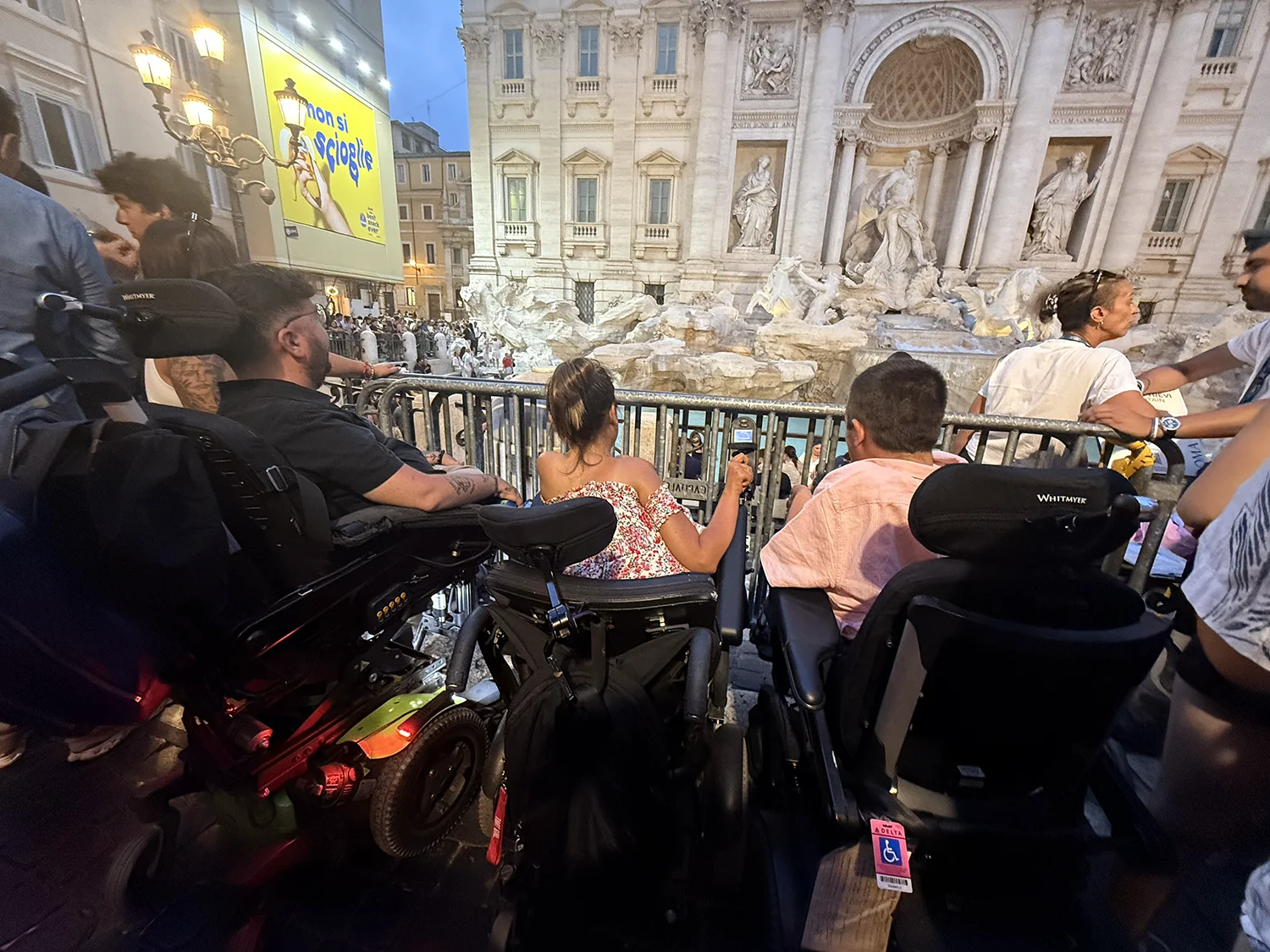 Three people in wheelchairs from behind looking toward the trevi fountain at night