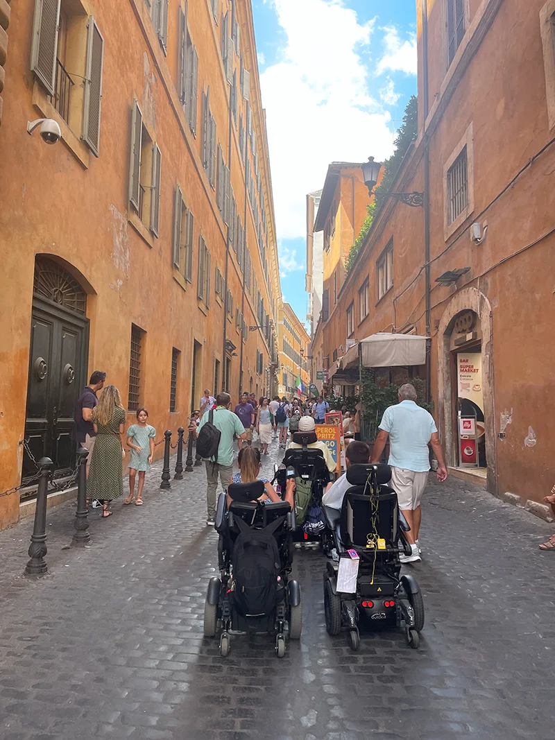 Three people in wheelchairs driving on cobblestones through an Italian street