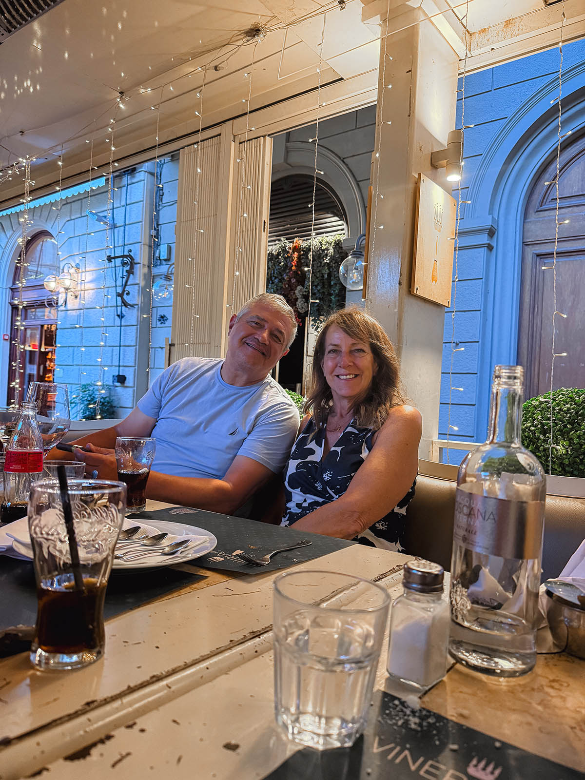 two people at a table smiling with empty plates and fairy lights behind them