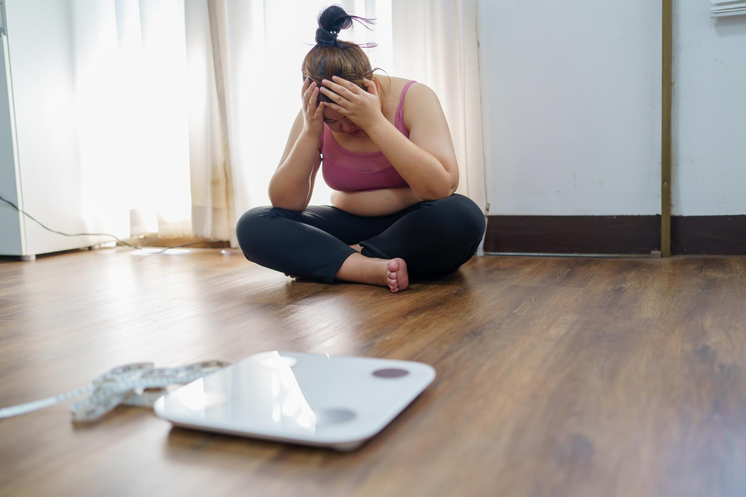 A woman sits on the floor, her hands to her head, looking stressed out. A scale is in the foreground.