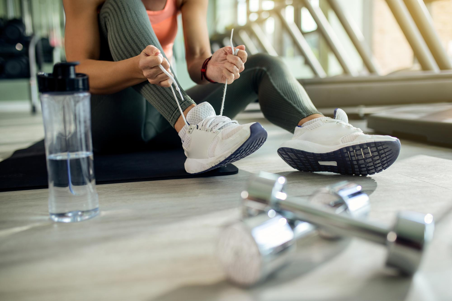 A woman sits on the floor of a gym, tying her shoe lace. Hand weights and a water bottle sit on the floor next to her.