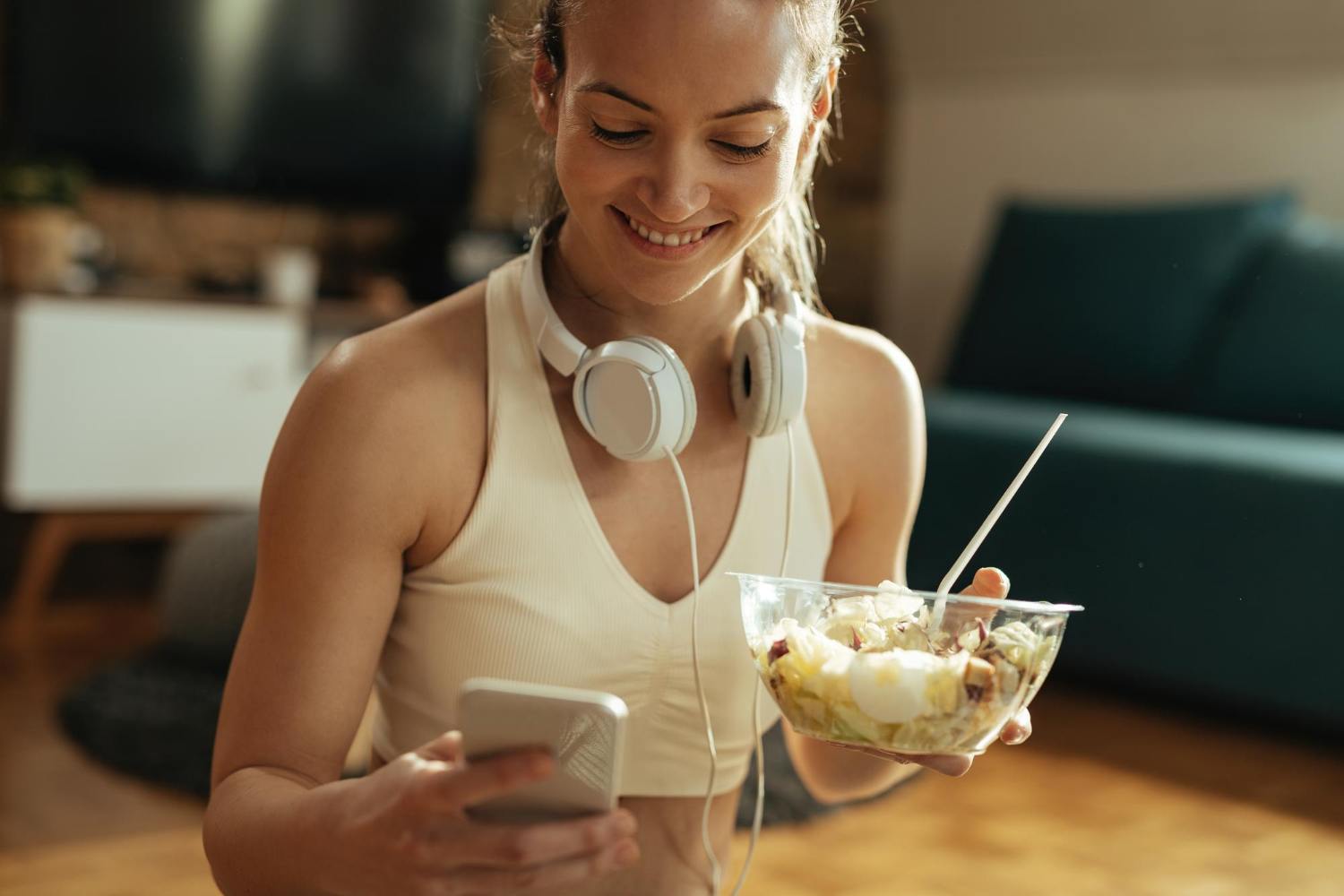 A woman enjoys a yogurt bowl with fruit after a workout. She's wearing workout gear, headphones, and is holding her smartphone.