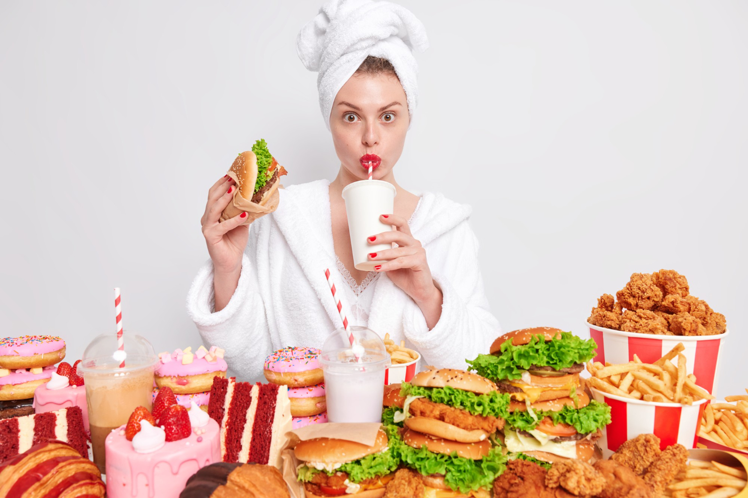 A woman in a bathrobe is pictured drinking a milkshake and holding a cheeseburger. The table in front of her is filled with junk food - burgers, fries,, cakes, and donuts.
