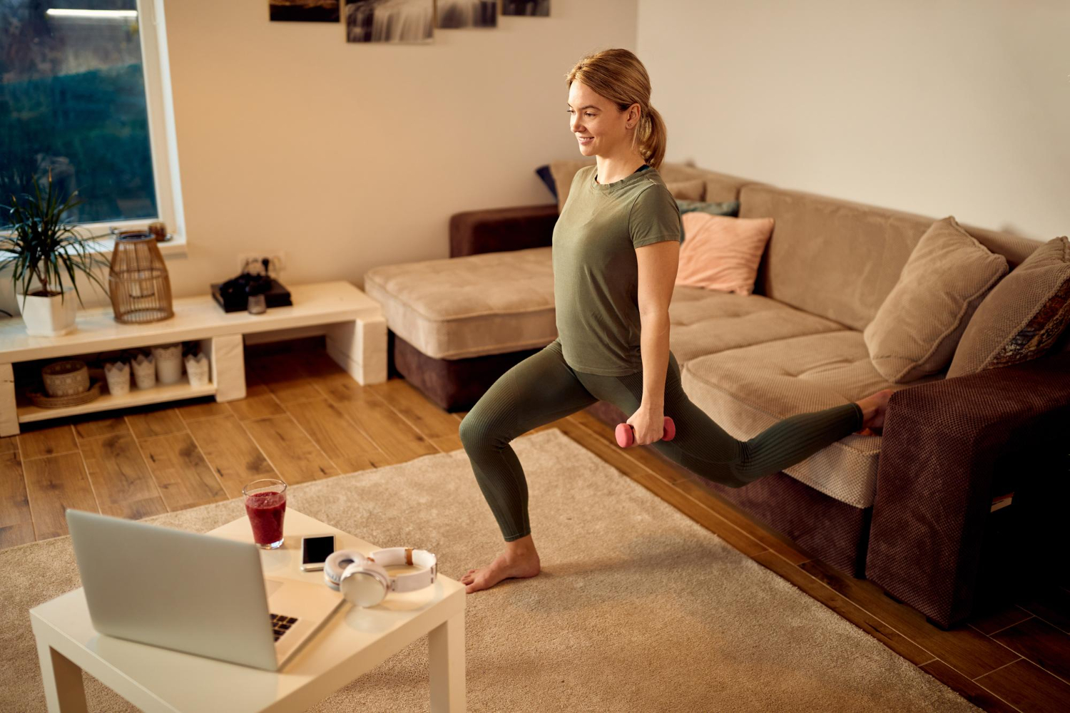 A woman works out in her living room, doing supported lunges on her couch and holding small weights.