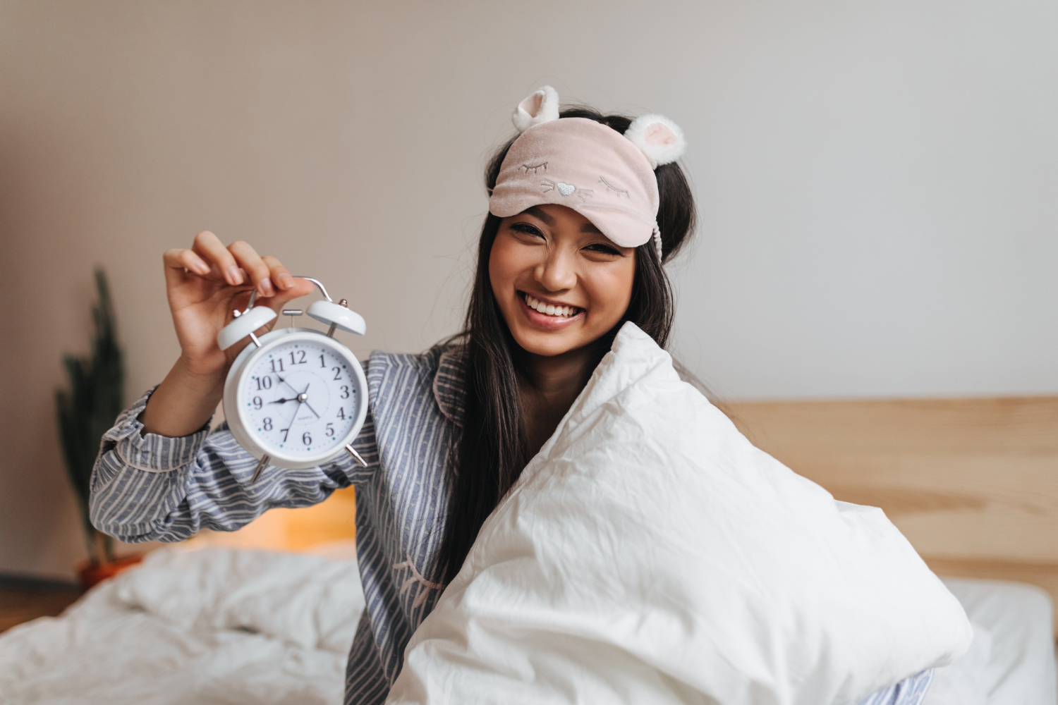 A young woman wakes up in bed, wrapped in her covers, wearing an eye mask and holding an alarm clock.