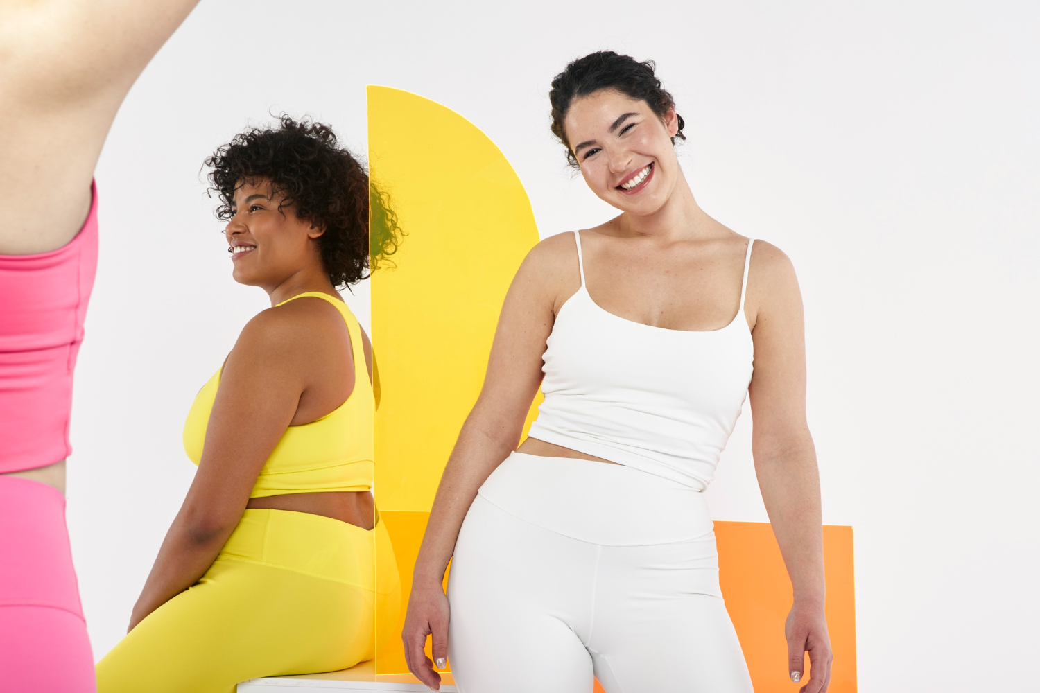 Two women standing next to each other, facing opposite directions. One is wearing a yellow workout set and the other a white workout set.