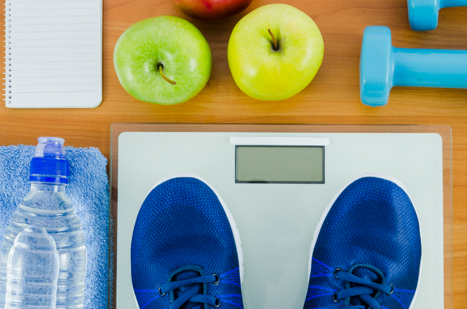 A close up image of a person's feet standing on a scale, surrounded by small weights, apples, and a bottle of water