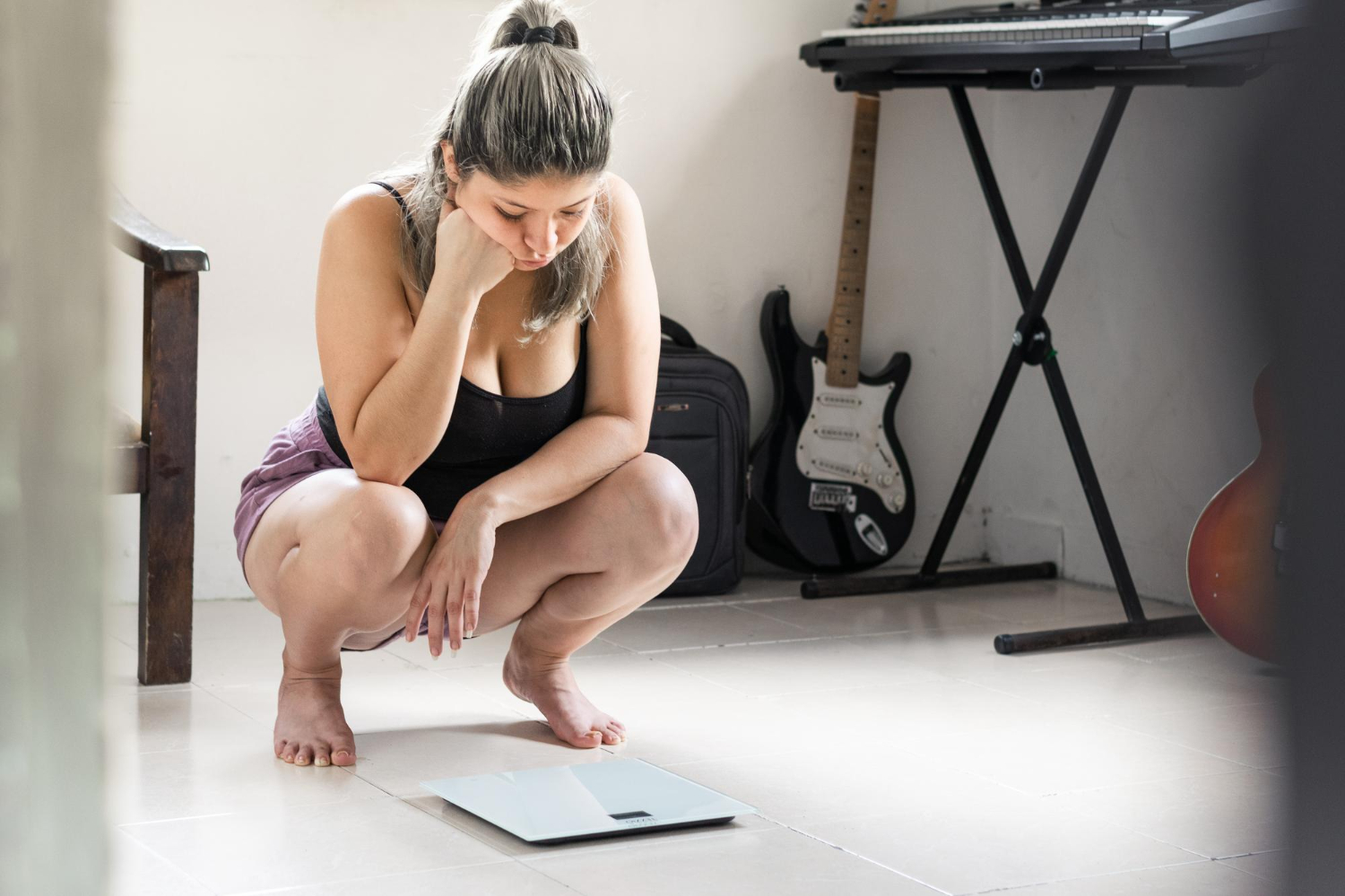 A woman is seen squatting down next to a scale, her head resting in her hands, looking frustrated. 