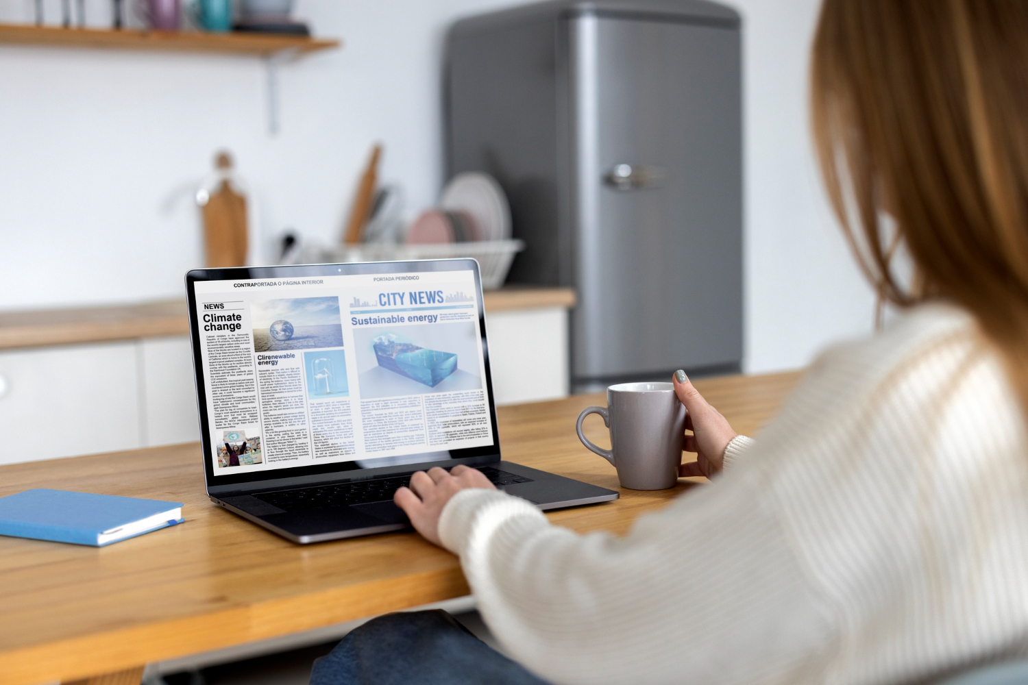 A woman sitting at a kitchen table, holding a cup of coffee and looking through news headlines on a laptop computer.