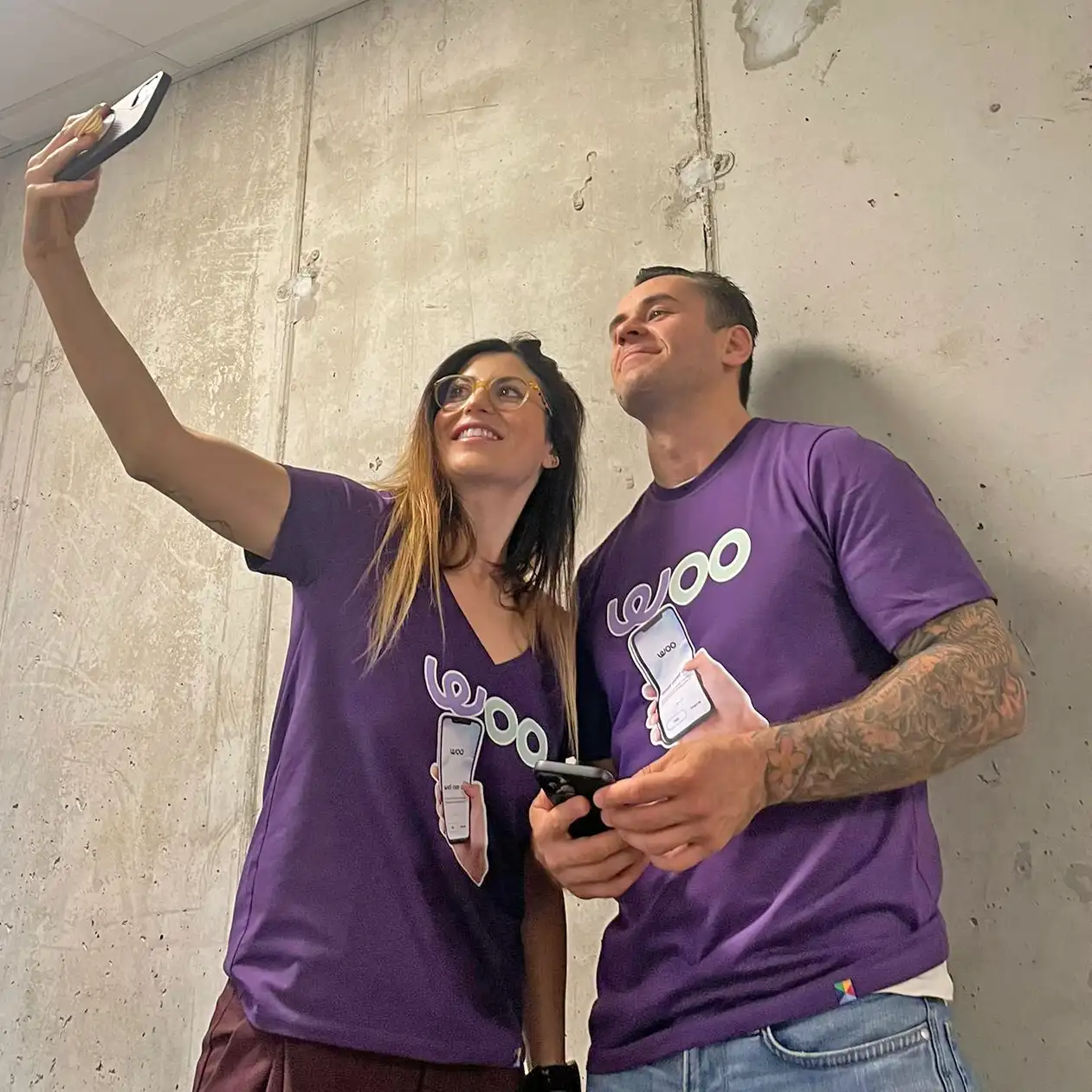 Dwoje młodych ludzi w fioletowych koszulkach robiących selfie na tle betonu.