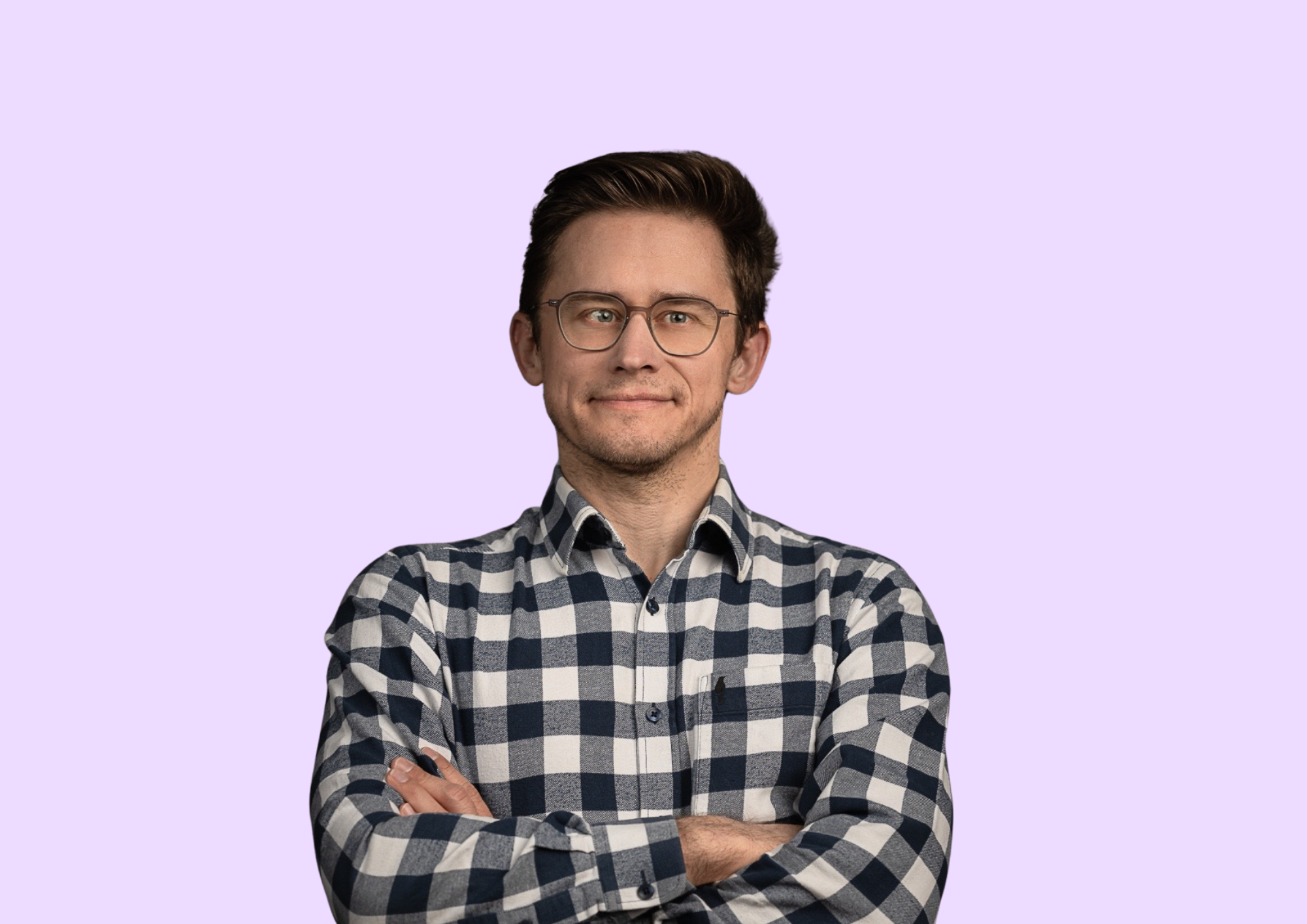 Mężczyzna w okularach w koszuli w kratę z rękami skrzyżowanymi na klatce piersiowej na jasnofioletowym tle.