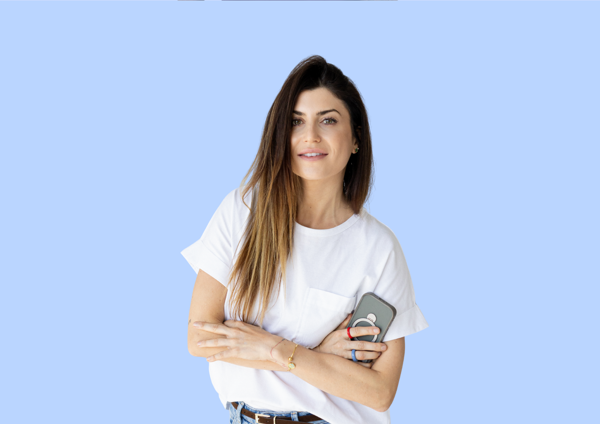 Uśmiechnięta kobieta z długimi brązowymi włosami, ubrana w białą koszulkę, trzymająca telefon z etui na jasnoniebieskim tle.