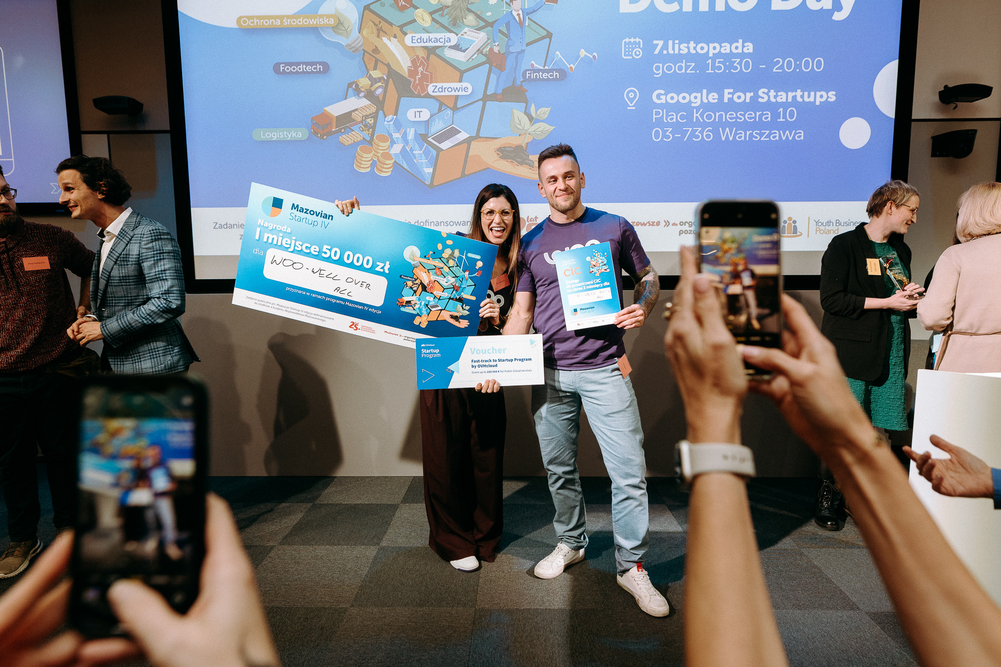 Para na scenie trzymająca duży czek na 50 000 zł za pierwsze miejsce oraz voucher, podczas ceremonii wręczenia nagród Startupów Mazowieckich.