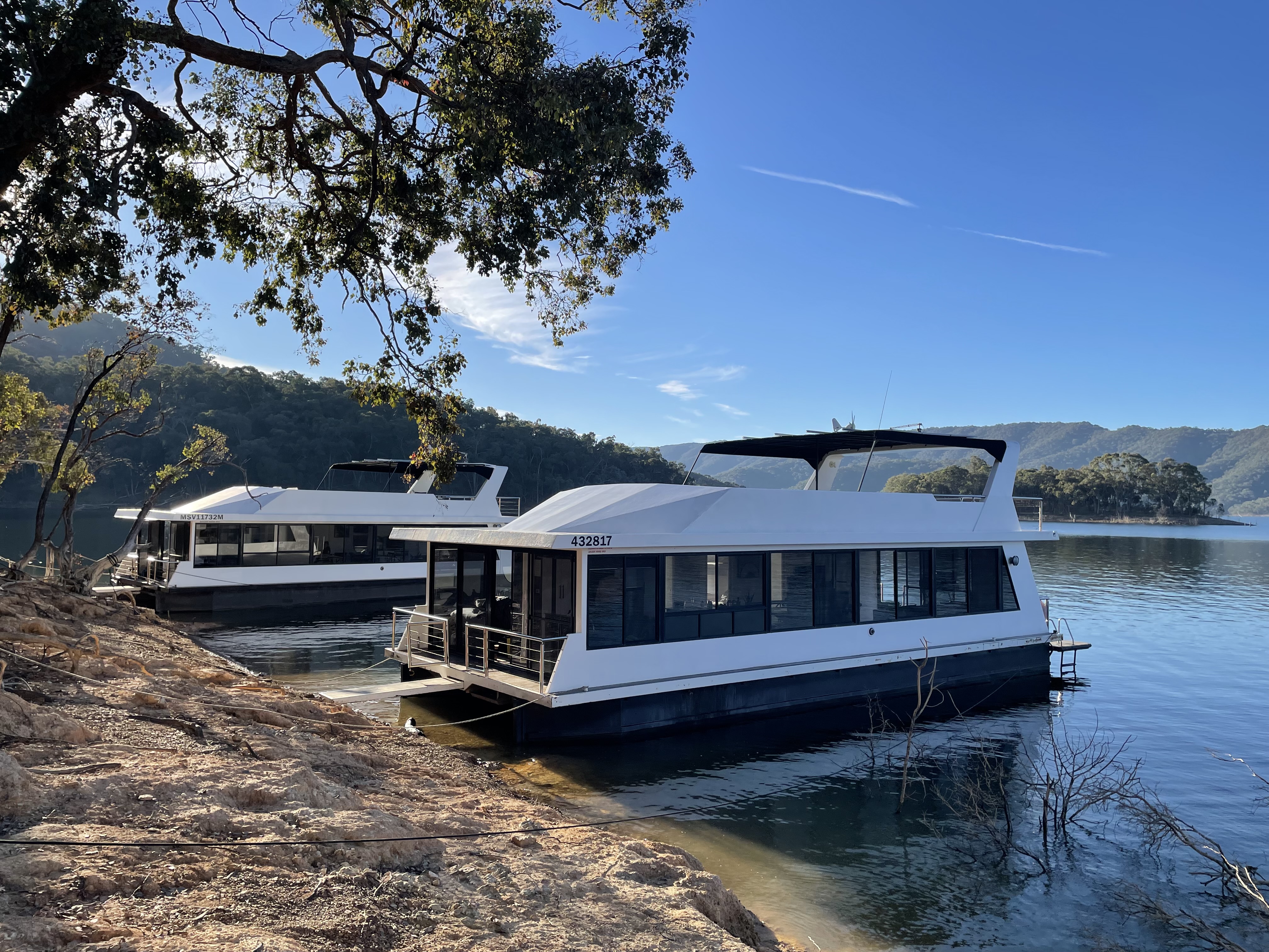 Two white houseboats moored at a tree-lined rocky shore, with forested hills and calm water in the background.
