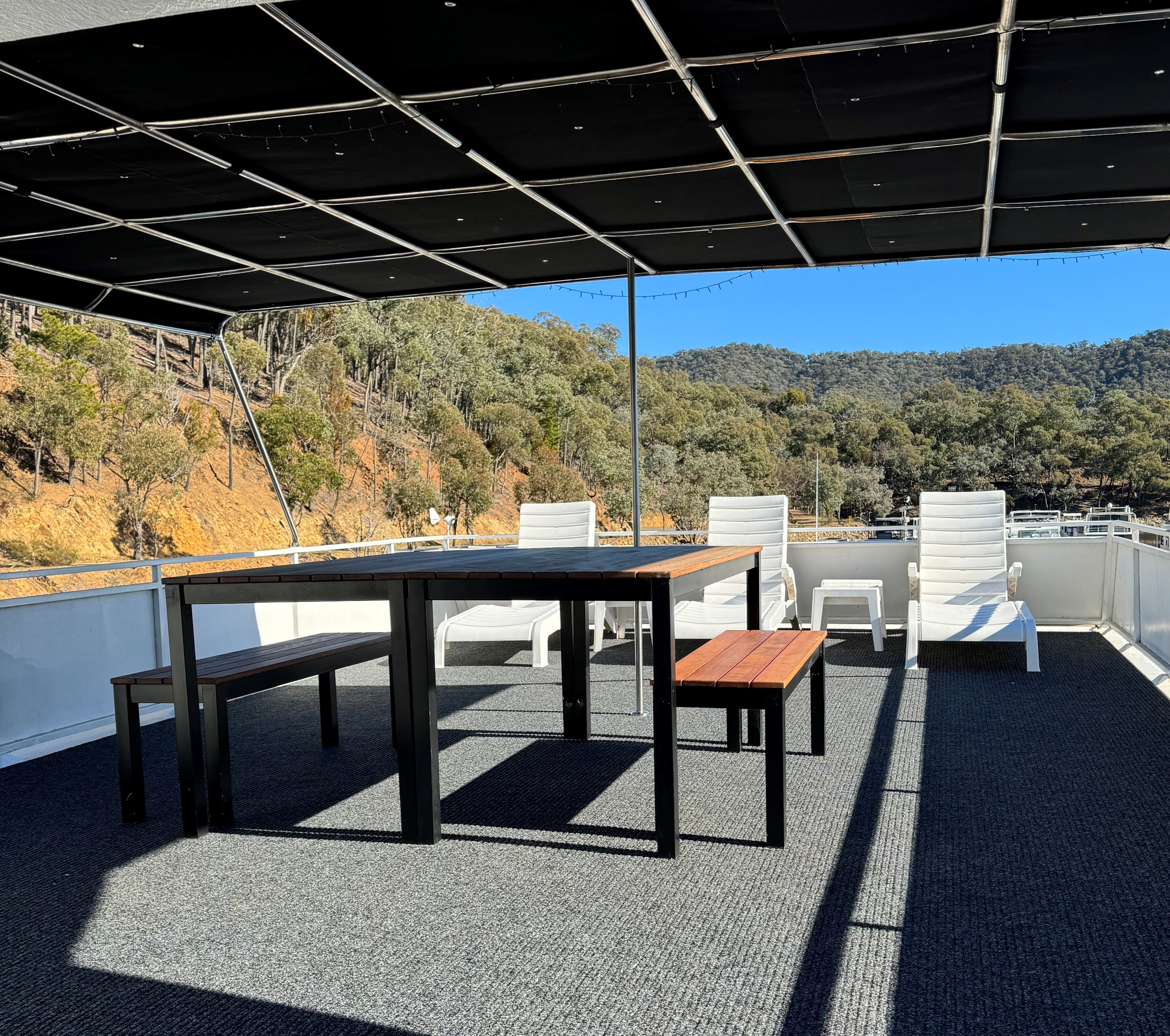 Rooftop entertaining deck with a wooden dining table, benches and white reclining chairs under a shade canopy with fairy lights.