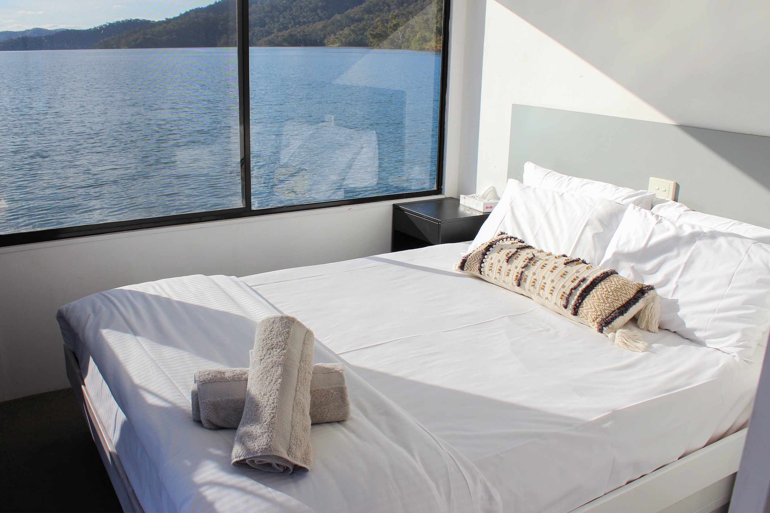 Bedroom with white bedding, a patterned bolster cushion and rolled towels, large window overlooking the lake and hills.