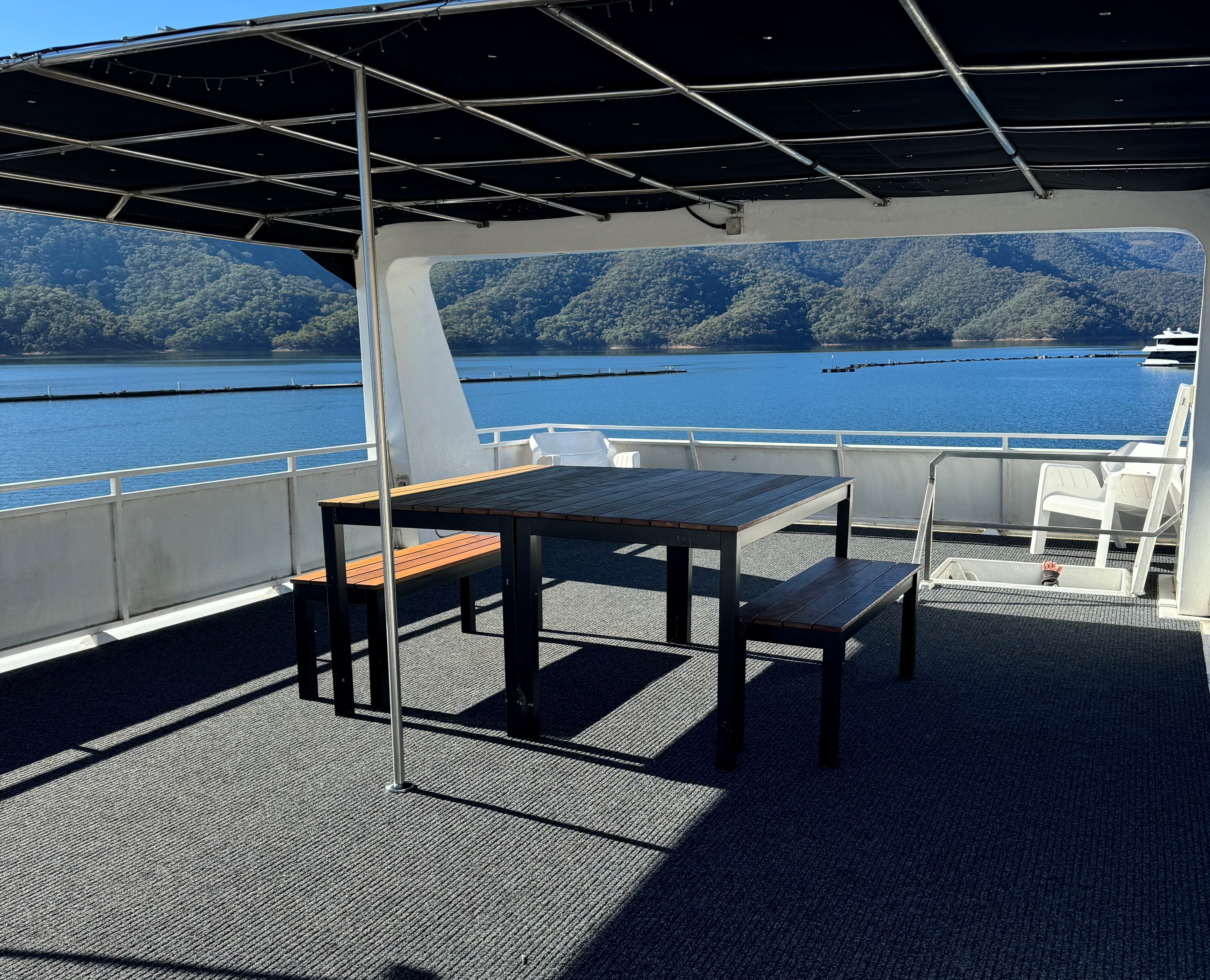 Covered rooftop deck with a dark timber-effect outdoor table and benches, white lounge chairs, overlooking the marina and lake.