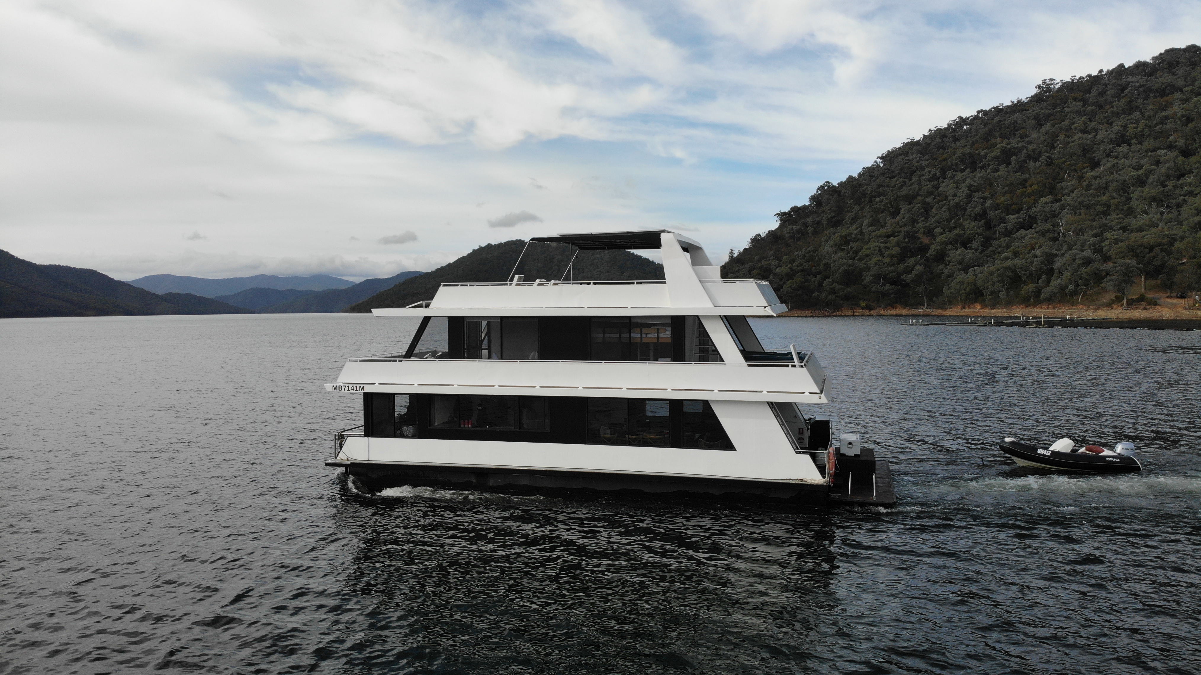 The Diplomat houseboat underway on the lake with a small dinghy following alongside, hills in the background.