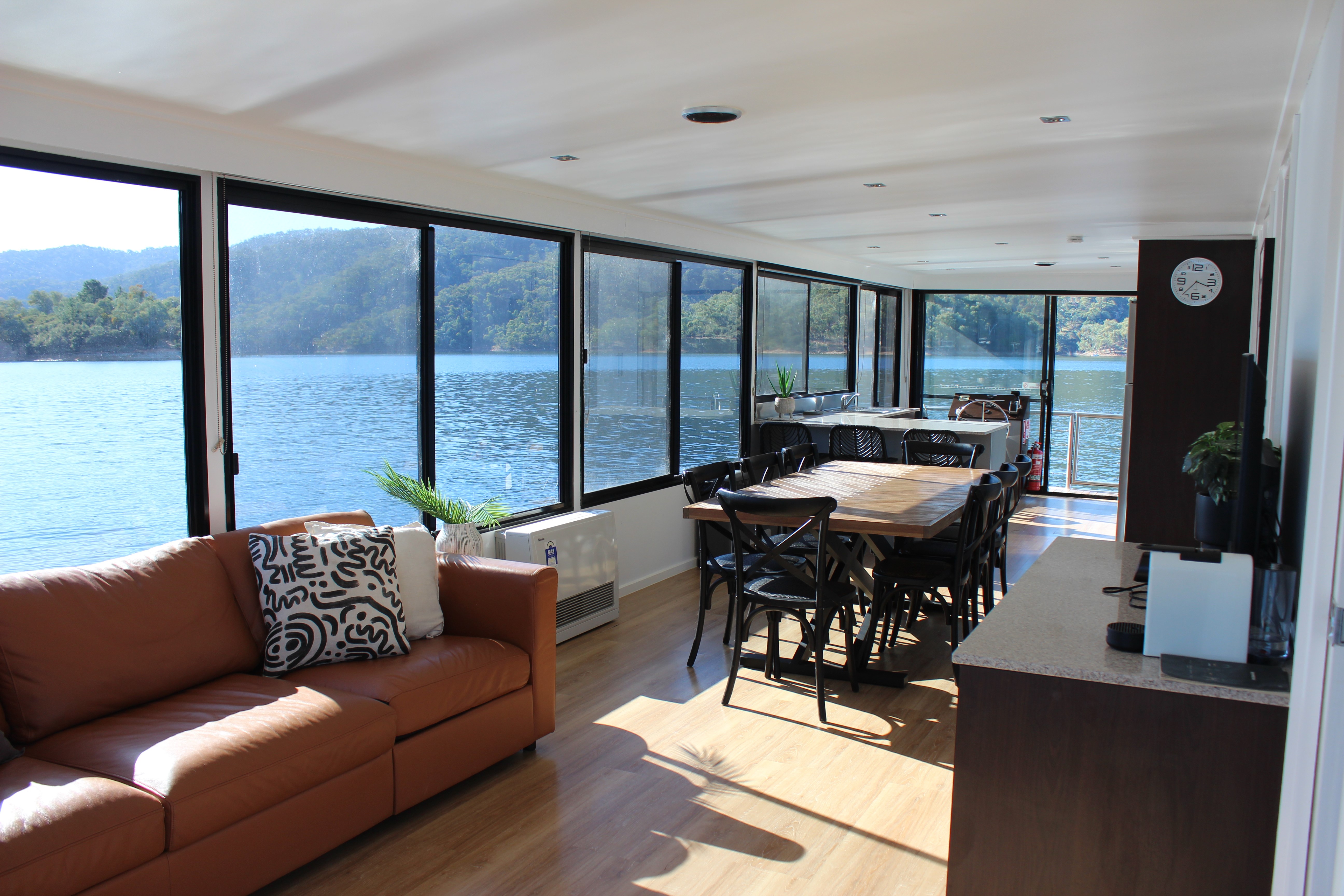 Open-plan living area with an orange leather sofa, patterned cushions, a wooden dining table with black chairs, and a wall of windows overlooking the lake.