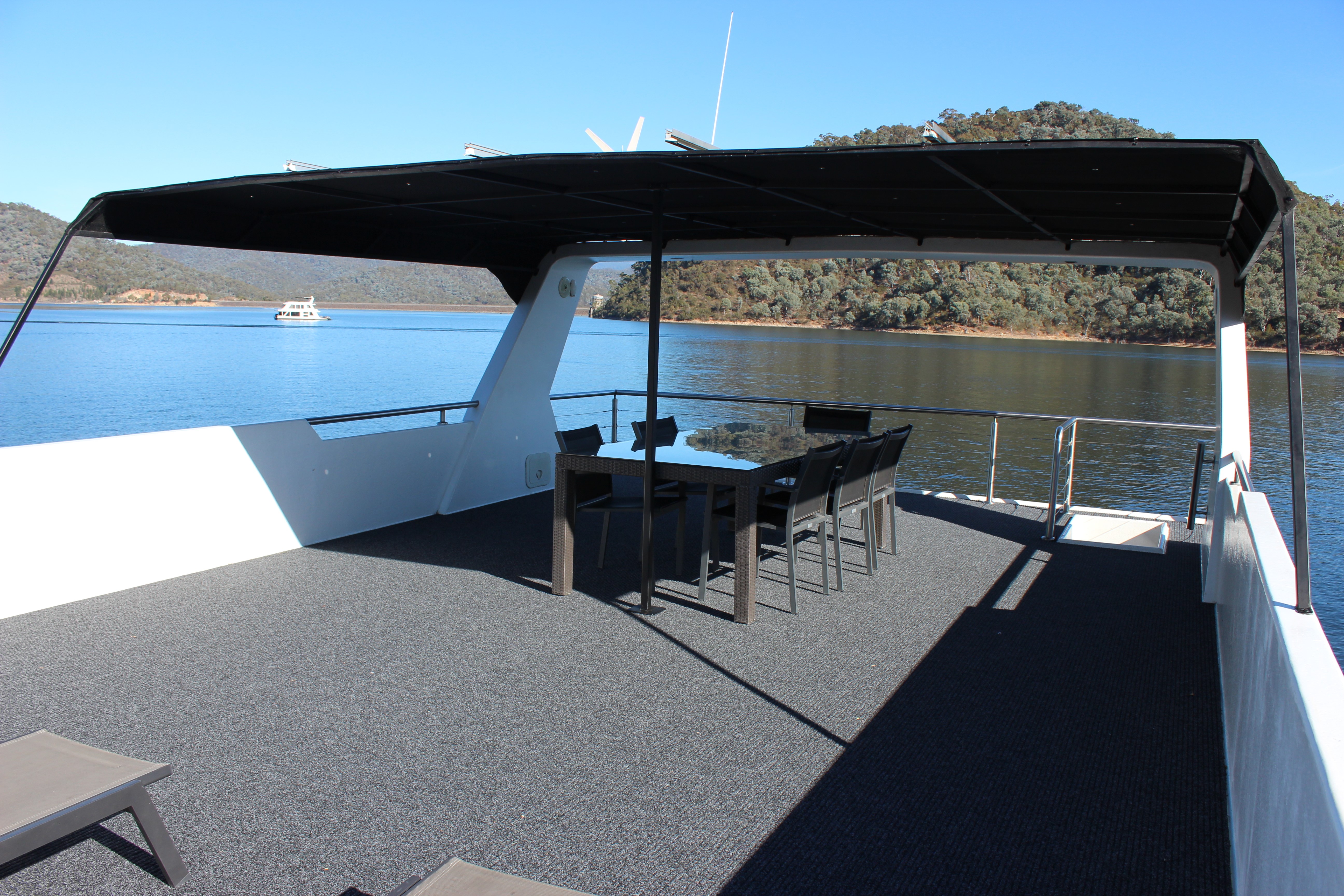 Covered rooftop deck with a black dining table and chairs, sun lounges and solar panels on the shade canopy, overlooking the lake and hills.