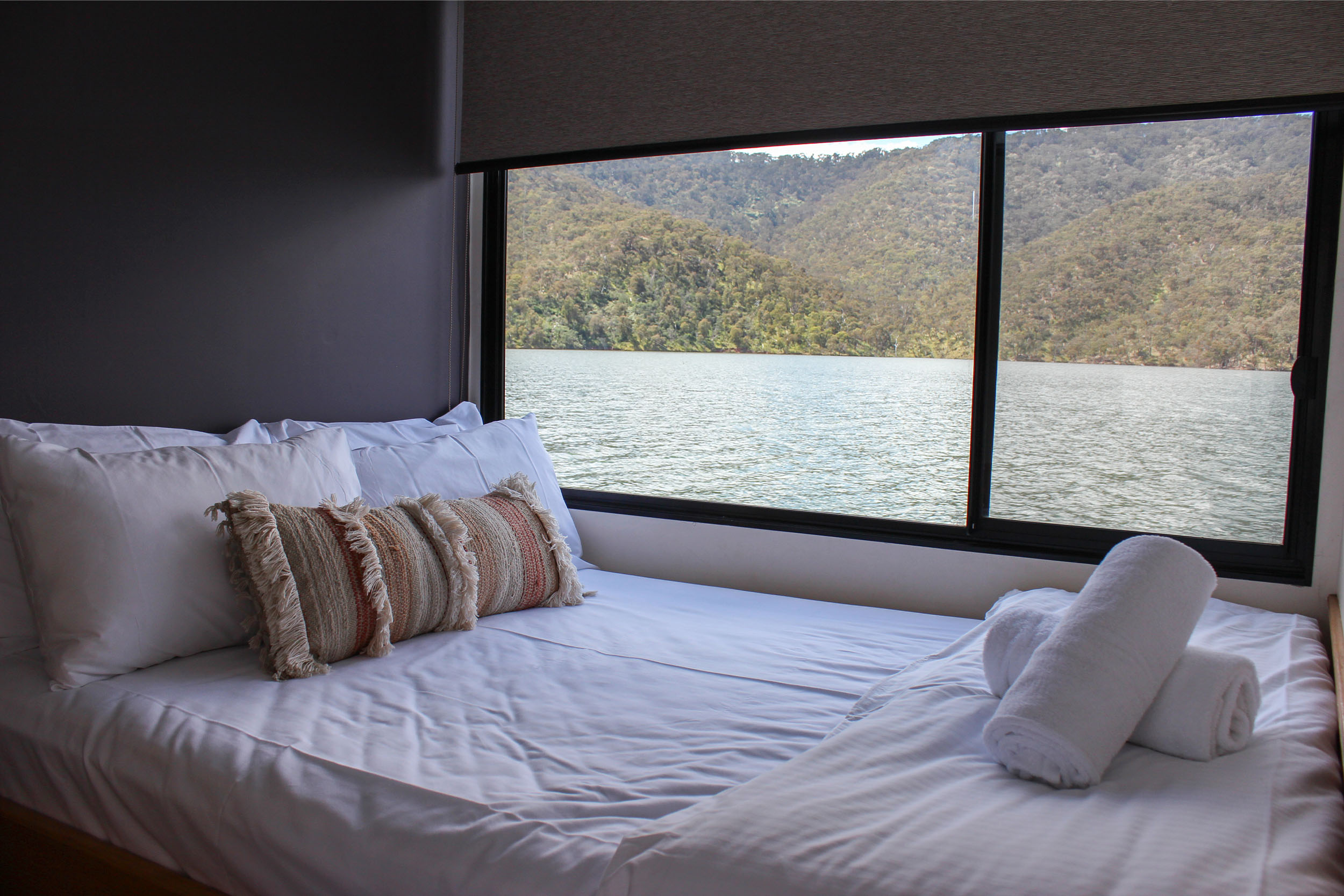 Queen bedroom with white bedding, a patterned bolster cushion and rolled towels, large window overlooking the lake and hills.