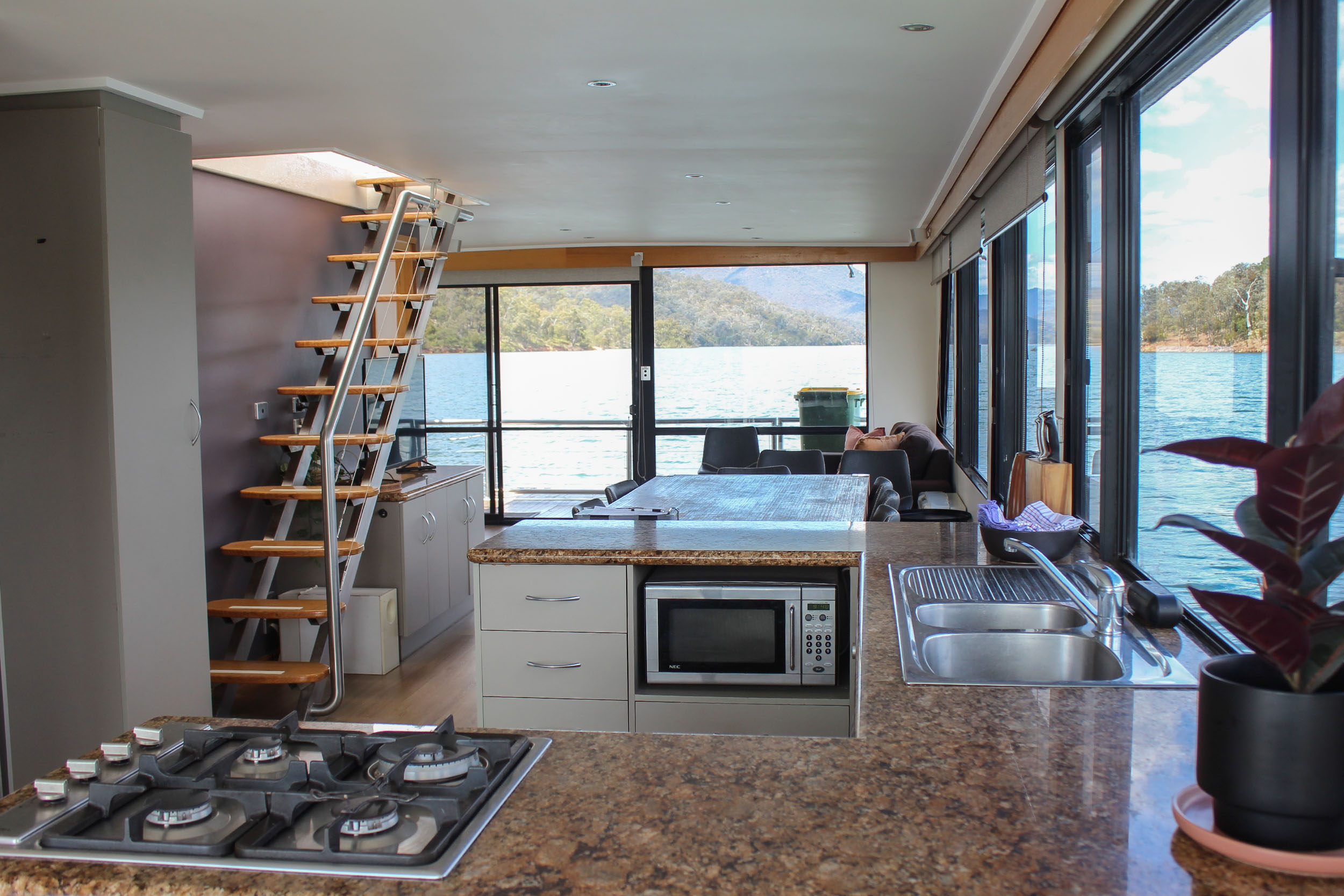 Kitchen area with a gas cooktop, granite benchtop, microwave and stainless sink, internal staircase, lounge and lake view beyond.