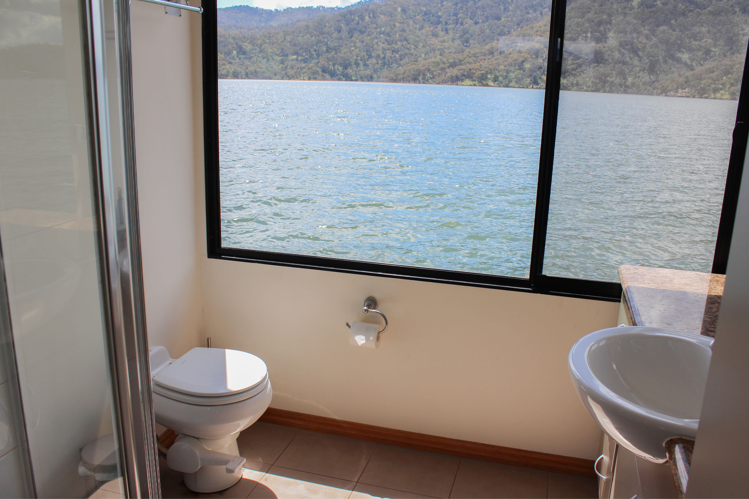 Bathroom with a toilet and pedestal-style sink, tiled floor, window overlooking the lake and hills.