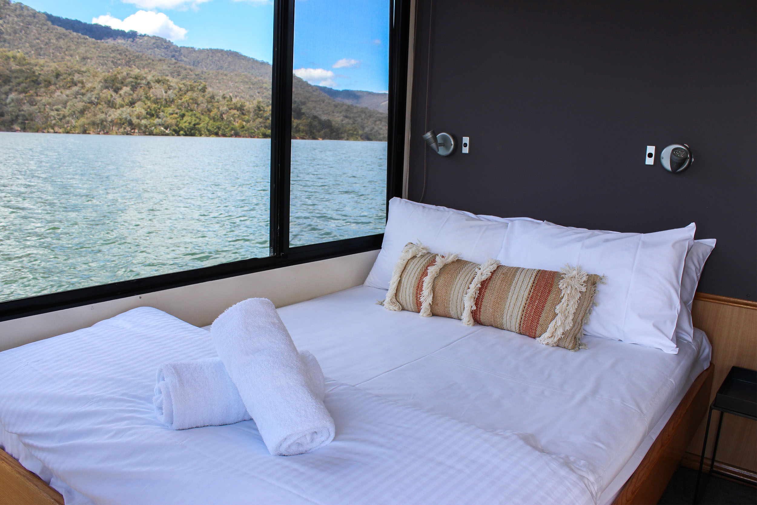 Queen bedroom with a dark feature wall, patterned bolster cushion, white bedding and rolled towels, lake and hills view through the window.