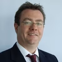 Professional headshot of Alan Wilkinson wearing a dark suit, white shirt, and red tie.