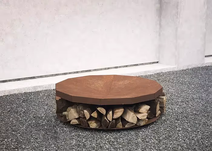 Braciere moderno da esterno con piano superiore ottagonale effetto corten e vano inferiore aperto contenente legna da ardere, posizionato su ghiaia grigia.