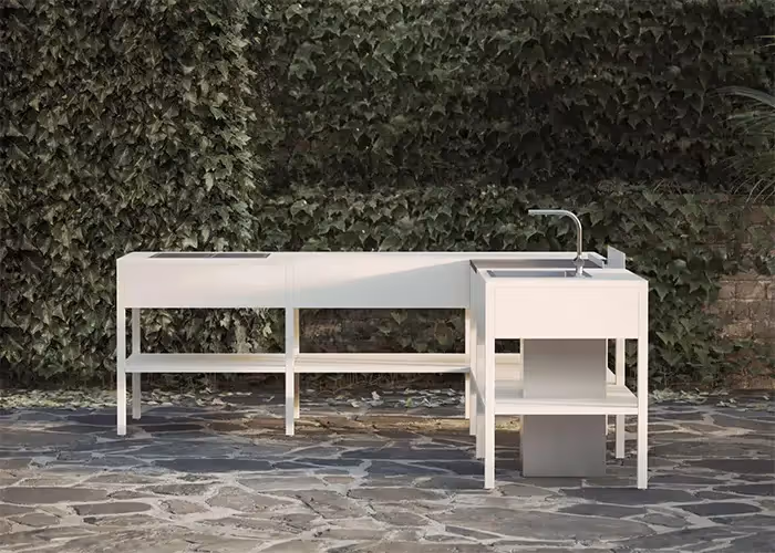 Cucina da esterno moderna in metallo bianco con lavello integrato e ampio piano di lavoro, posizionata su una terrazza in pietra davanti a una parete ricoperta di edera.