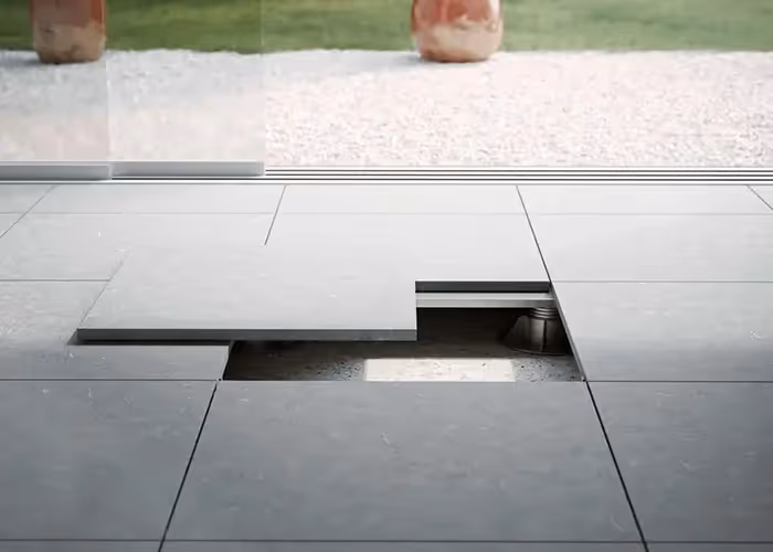 Pavimentazione moderna da esterno con piastrelle grigie modulari e pannello ispezionabile sollevato, davanti a una vetrata.