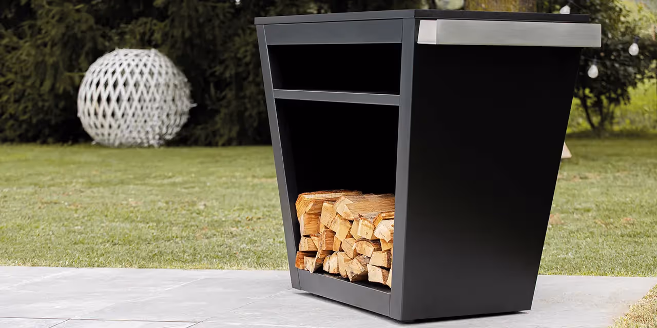 Banco barbecue moderno da esterno in metallo nero con vano inferiore porta legna e piano laterale in acciaio, posizionato in un giardino.