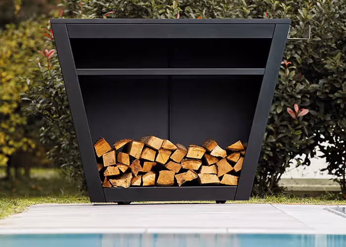 Banco barbecue da esterno con struttura geometrica in metallo nero e vano inferiore riempito con legna da ardere, accanto a una piscina.