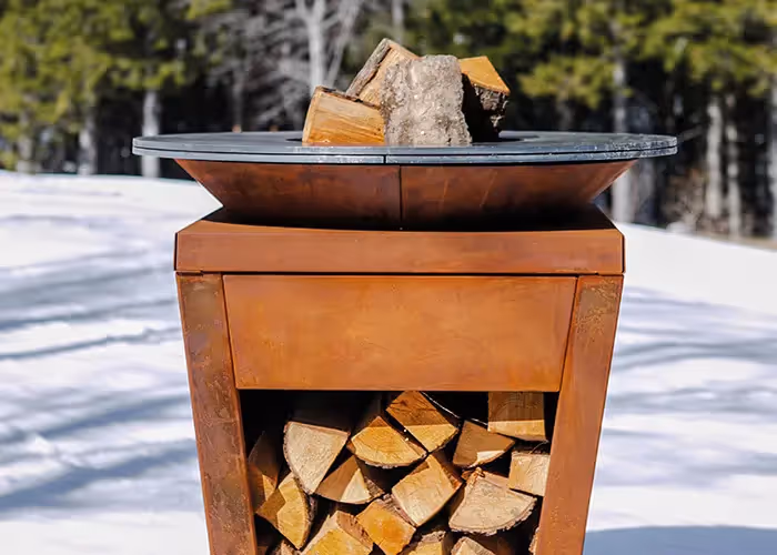 Barbecue circolare in acciaio corten color ruggine con legna da ardere, posizionato in un paesaggio innevato.