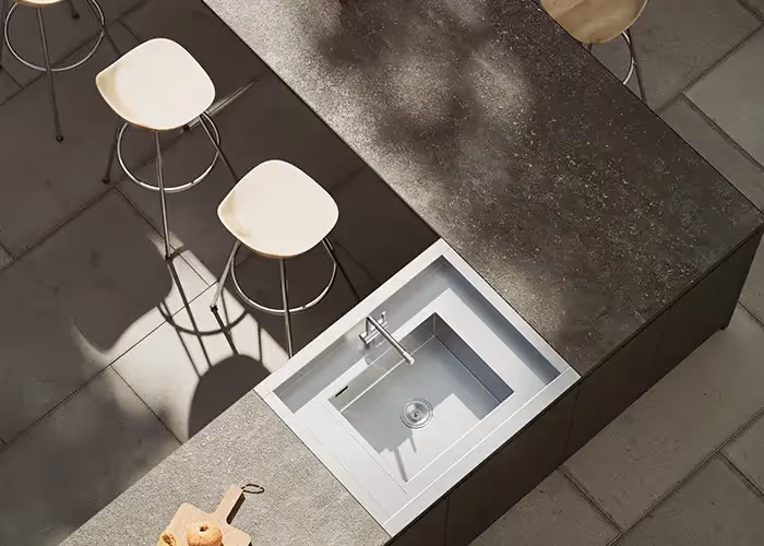 Vista dall’alto di una cucina da esterno moderna con piano effetto pietra, lavello integrato e sgabelli chiari su pavimentazione in pietra.