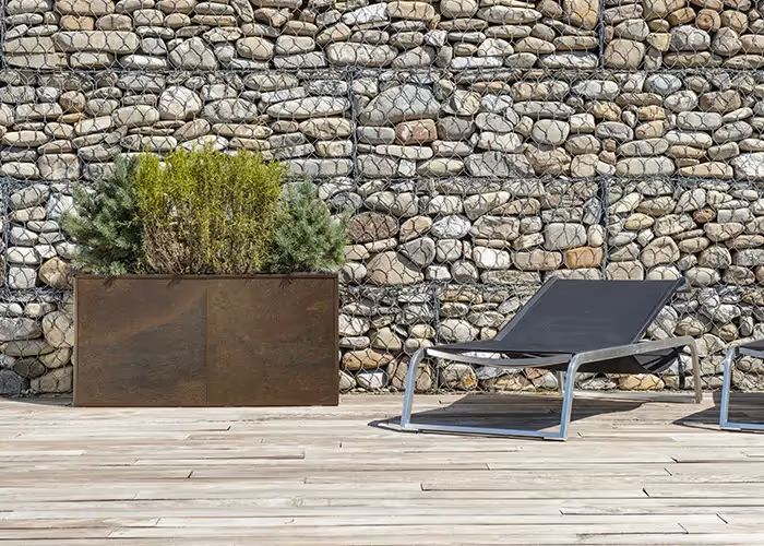 Fioriera rettangolare effetto corten con arbusti ornamentali, posizionata su una terrazza in legno accanto a lettini da esterno.