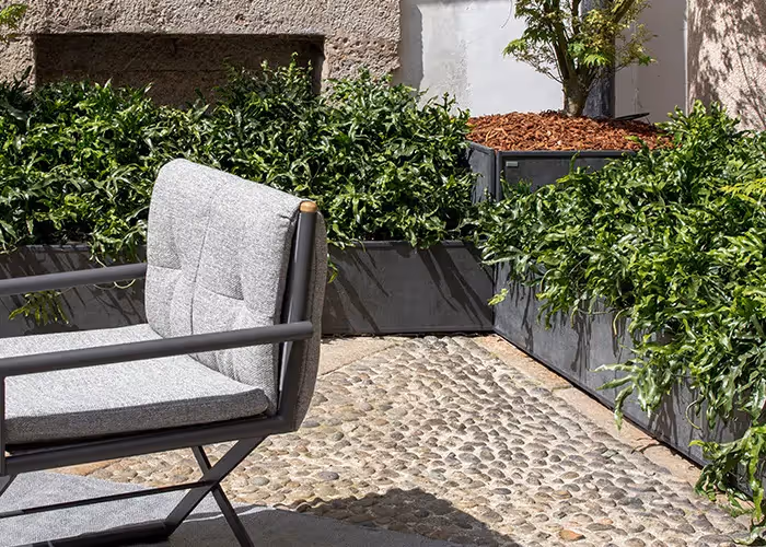 Area lounge da esterno con poltrona imbottita grigia e lunghe fioriere modulari scure contenenti vegetazione verde.