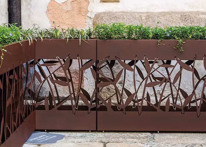 Fioriere modulari effetto corten con pannelli decorativi traforati e piante ornamentali, posizionate in un cortile esterno.