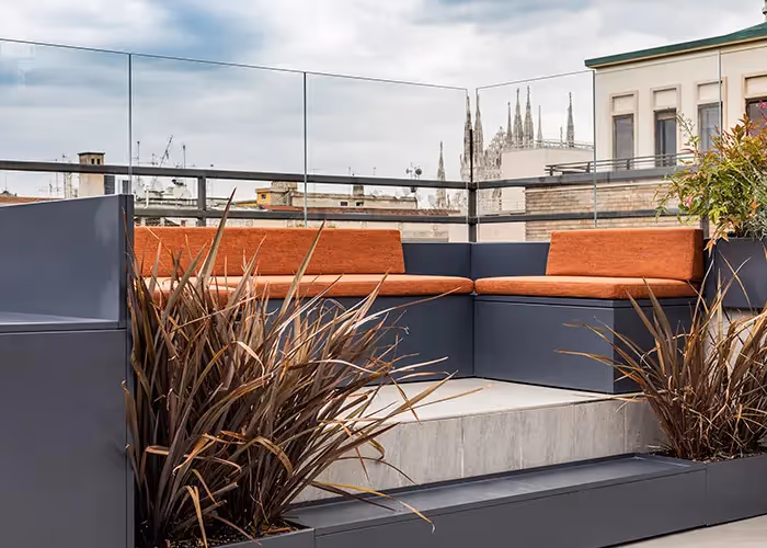 Area lounge moderna su terrazza urbana con sedute modulari da esterno in metallo scuro, cuscini arancioni e fioriere integrate con vegetazione ornamentale.