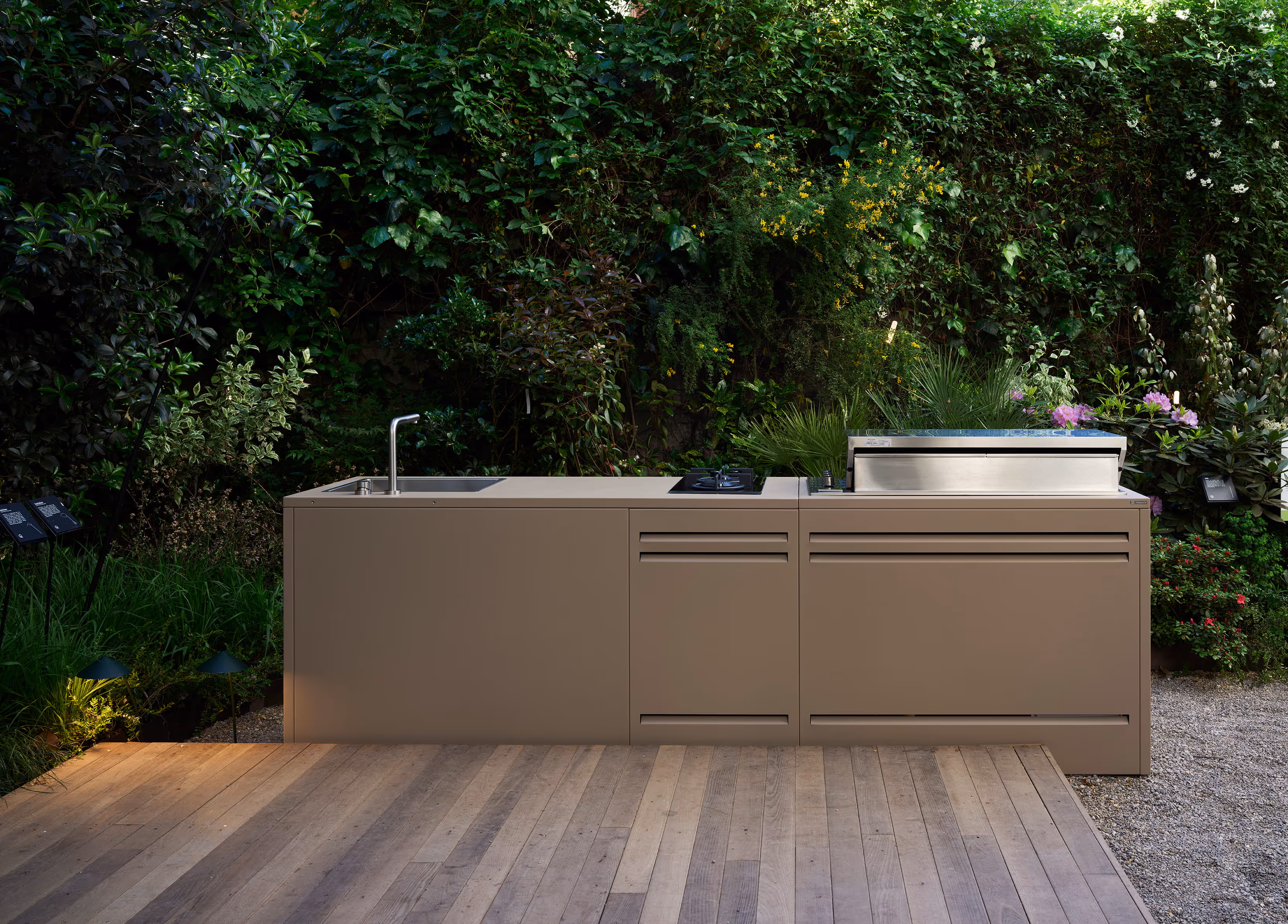 Cucina da esterno beige con lavello, fuochi e barbecue integrato, posizionata in un giardino lussureggiante.