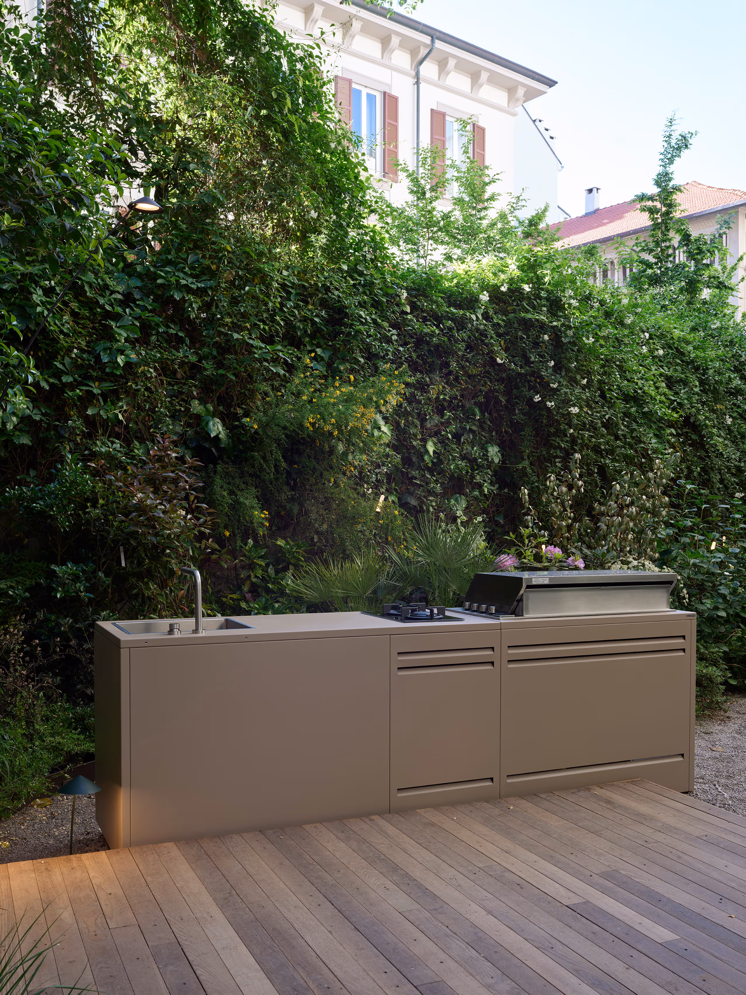 Cucina lineare da esterno installata su una pedana in legno in giardino, con siepe alta sullo sfondo.