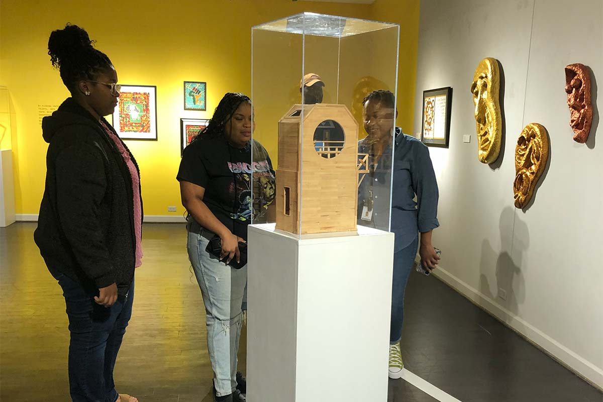 Two older Black teen girls look at an artwork under glass in an art gallery while their teacher, who is a Black woman, looks on.