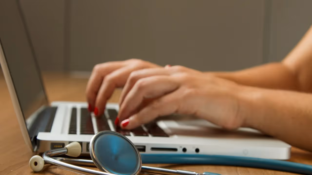 person typing on a laptop keyboard with a stethoscope resting nearby on the desk