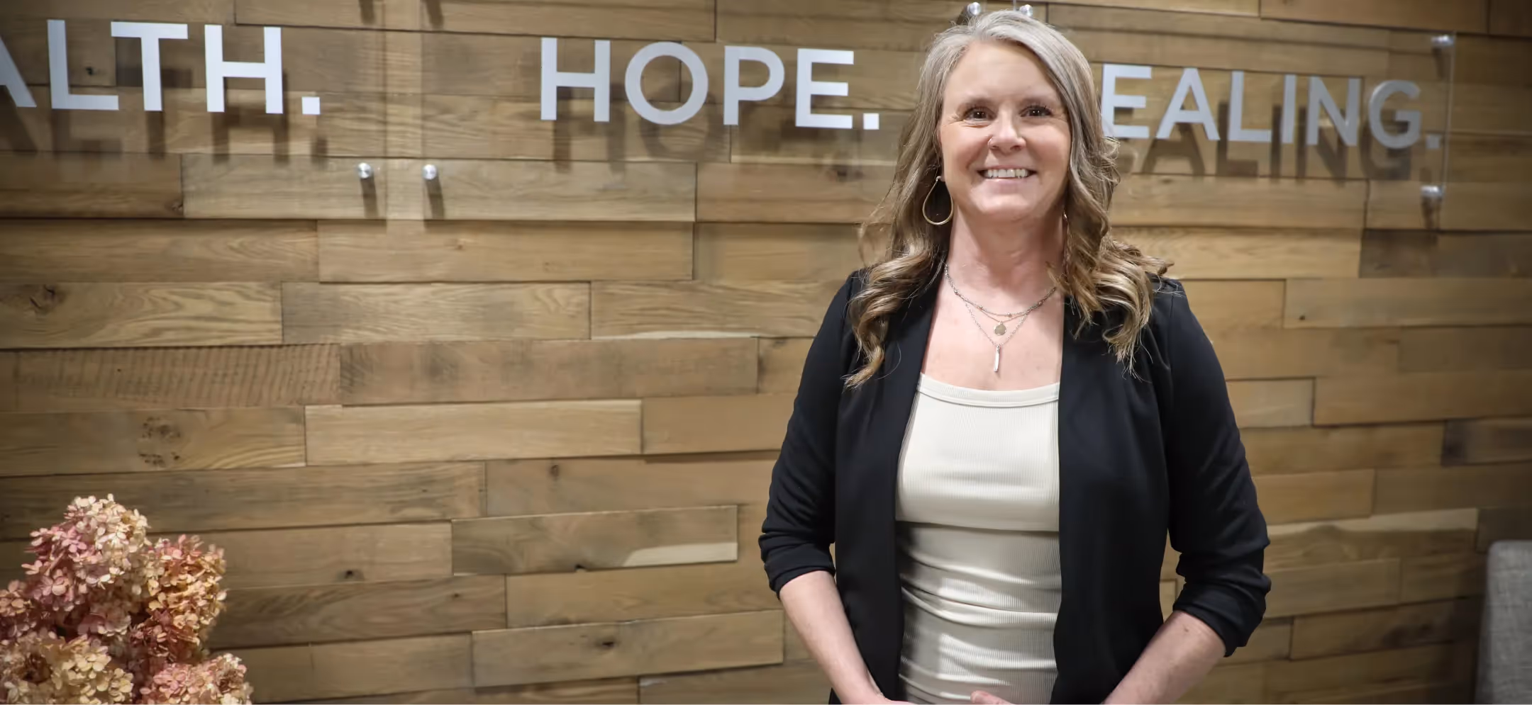 person standing in front of a wood‑paneled wall with the words “health hope healing” displayed behind them
