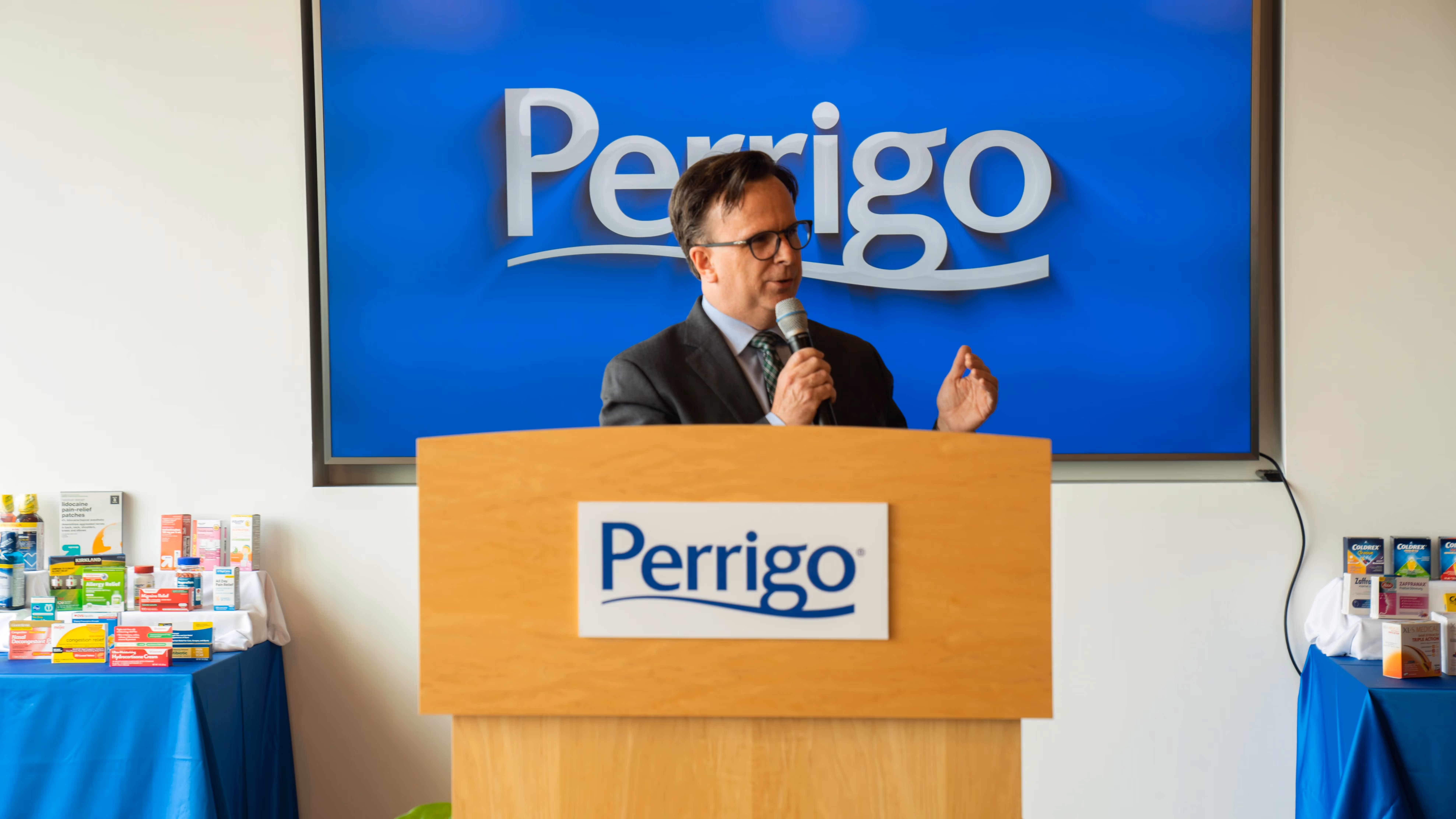speaker standing at a Perrigo podium during an event with product displays arranged on side tables
