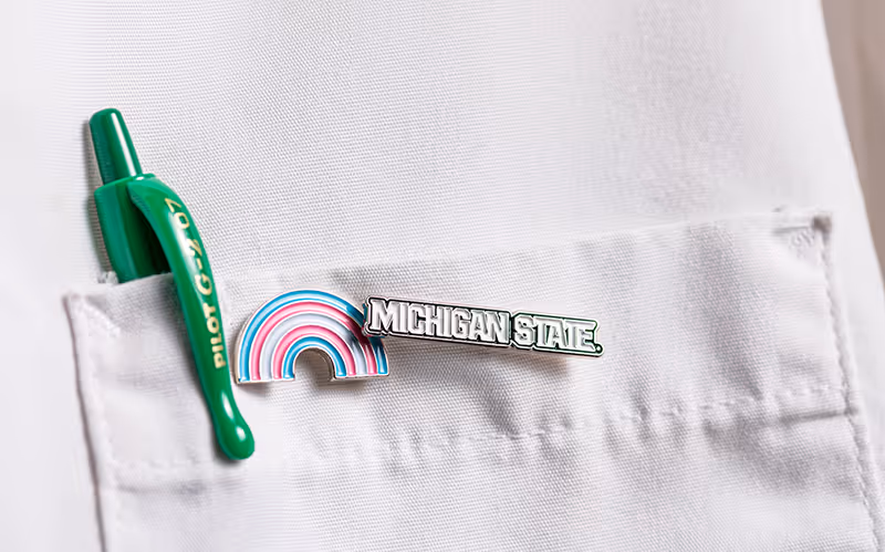 Close-up of a white lab coat pocket holding a green pen and two enamel pins—one rainbow pin and one reading “Michigan State”—attached to the pocket seam.
