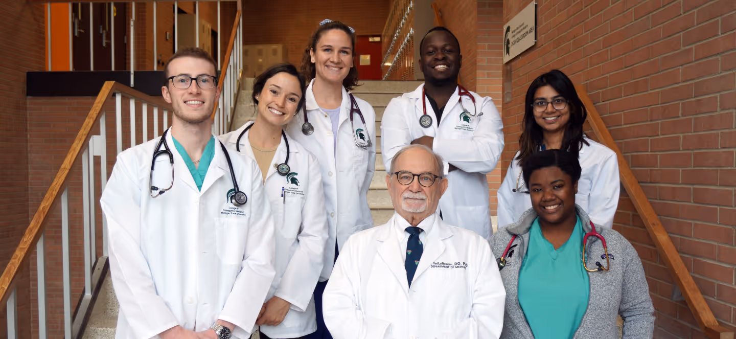 Group of medical students and faculty wearing white coats and stethoscopes pose together on a staircase inside a brick academic building.
