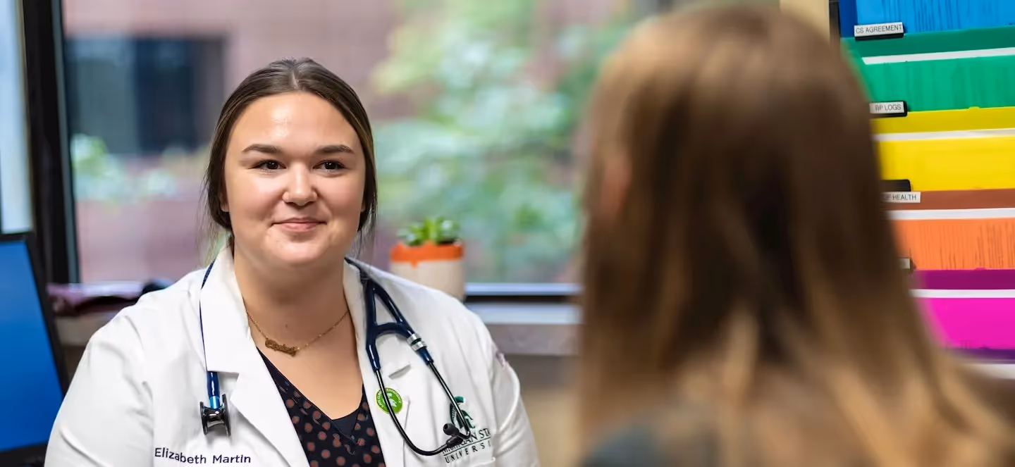 Health care provider in a white coat with a stethoscope smiles while speaking with a patient in a clinic exam room, with a window and organized medical files visible in the background.